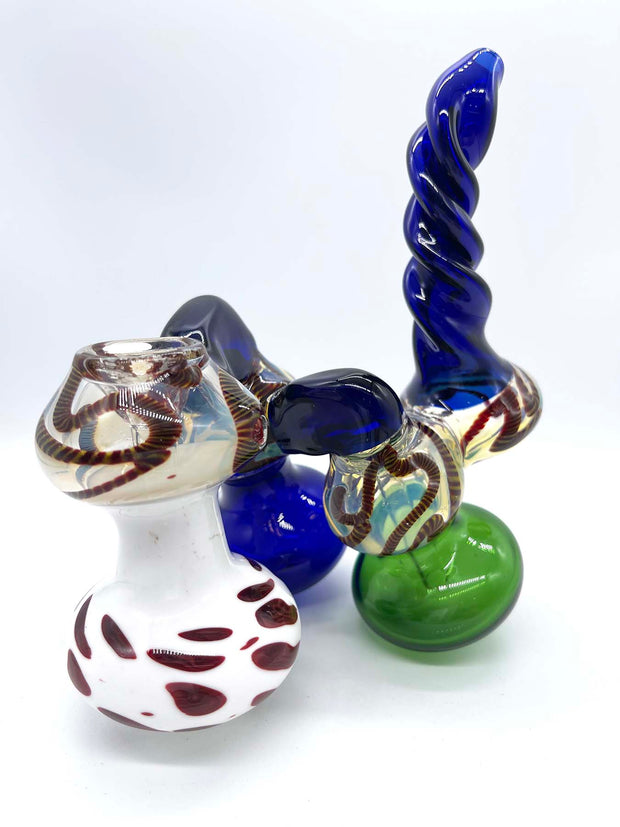 Photo of a three-chamber glass bubbler. The front chamber is colored white with smeared red dots, the middle chamber is blue, and the rear chamber is green. The bridges connecting each chamber and the twisted mouthpiece match the blue of the middle chamber. The bowl is blown into the top of the front chamber and the top of each chamber is fumed in silver and dark red rope.