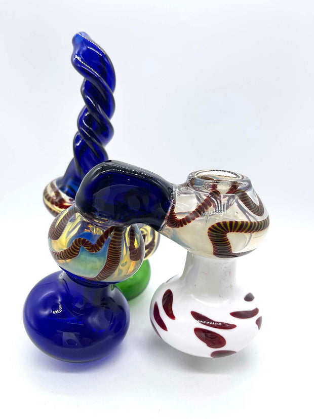 Side view photo of a three-chamber glass bubbler. The front chamber is colored white with smeared red dots, the middle chamber is blue, and the rear chamber is green. The bridges connecting each chamber and the twisted mouthpiece match the blue of the middle chamber. The bowl is blown into the top of the front chamber and the top of each chamber is fumed in silver and dark red rope.