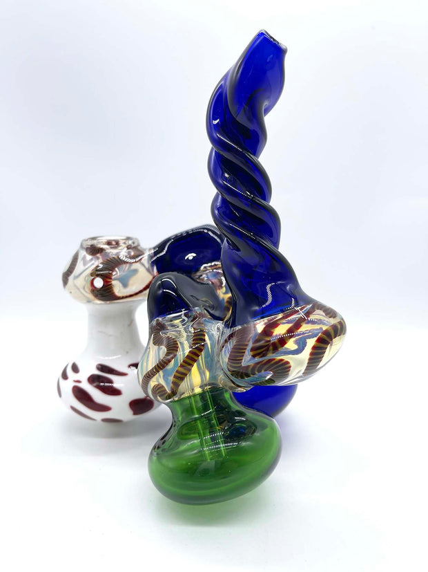 Rear view photo of a three-chamber glass bubbler. The front chamber is colored white with smeared red dots, the middle chamber is blue, and the rear chamber is green. The bridges connecting each chamber and the twisted mouthpiece match the blue of the middle chamber. The bowl is blown into the top of the front chamber and the top of each chamber is fumed in silver and dark red rope.